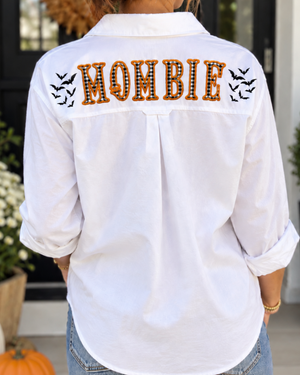 A woman viewed from behind wearing a white long-sleeve button-up shirt with 'MOMBIE' spelled out in orange applique letters on the back, flanked by black bat silhouettes. She is wearing blue jeans and standing outdoors in front of a house door with pumpkins and potted plants.
